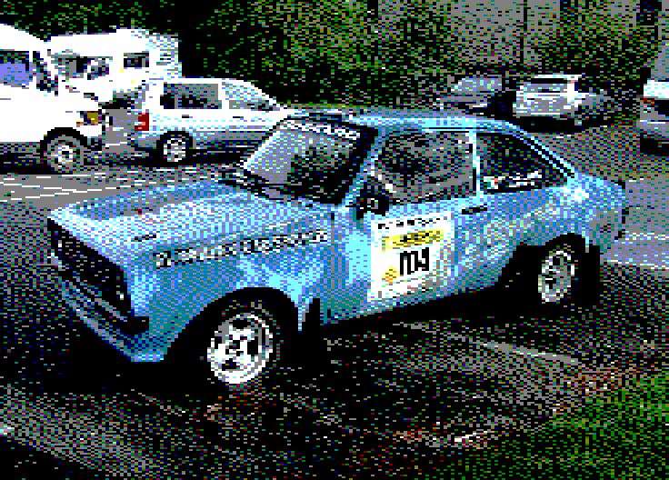 Image converted by 'CPiC'. Blue vintage rally Ford, with the number 104, parked in a parking lot with other cars and vans in the background.
