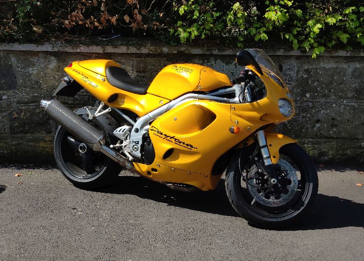 The yellow Triumph Daytona, sports motorcycle, featuring a sleek design, black seat, and silver exhaust pipe, parked on the side of an empty road in front of an ivy-covered stone wall.