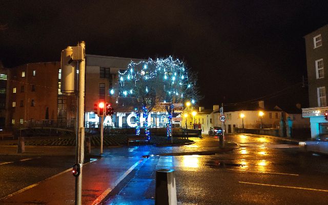 Night view of Carlow street with a large tree decorated with blue lights in the center. Traffic lights on the left and a building on the right.