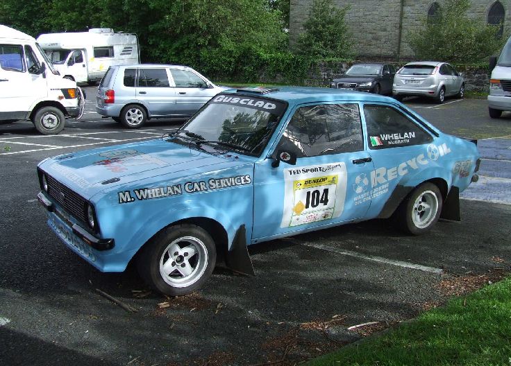 Blue vintage rally Ford, with the number 104, parked in a parking lot with other cars and vans in the background.