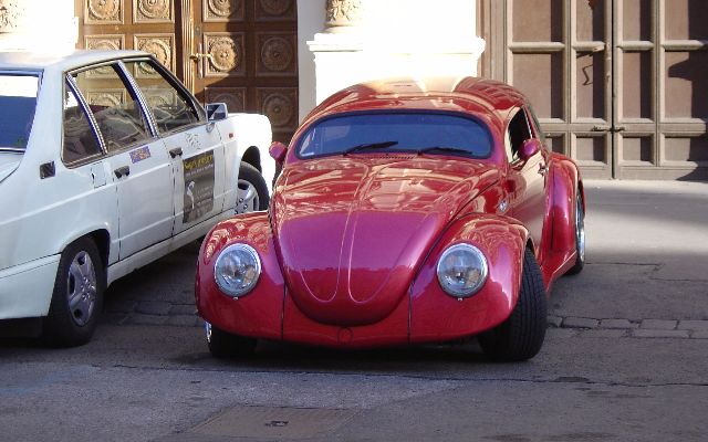 A classic, red, shiny Volkswagen Beetle, in excellent condition, parked on the street.