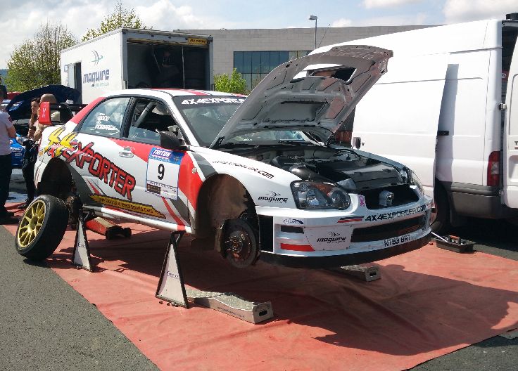 The white Subaru Impreza, rally car, with red and yellow accents and the number 9, on a red platform with its hood open. Other cars and equipment in the background.