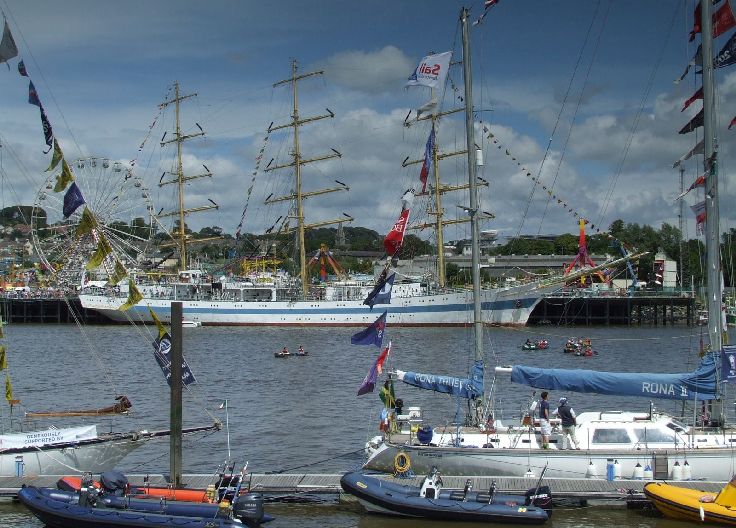 The harbor with several boats and ships moored, in the center a large white yacht with three masts. A Ferris wheel is visible in the background, and smaller boats with colorful flags to the right.