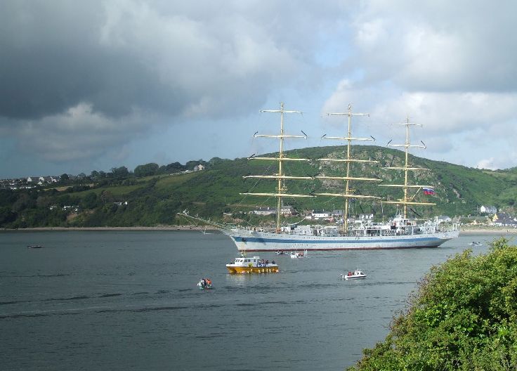 A large, blue and white, three-masted yacht surrounded by smaller boats on calm water. In the background, green hills with trees and houses under a cloudy sky.